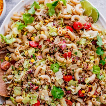 A platter of taco pasta salad with macaroni, ground meat, lettuce, tomatoes, corn, red onion, cilantro, and lime wedges.