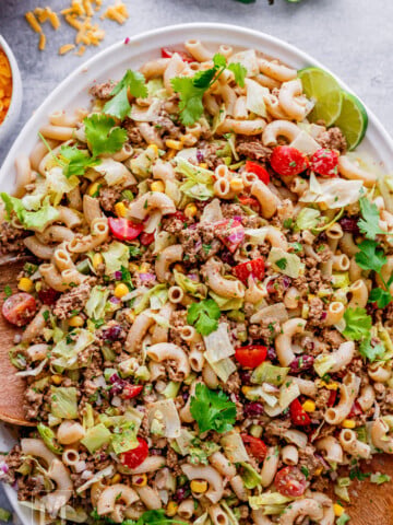 A platter of taco pasta salad with macaroni, ground meat, lettuce, tomatoes, corn, red onion, cilantro, and lime wedges.