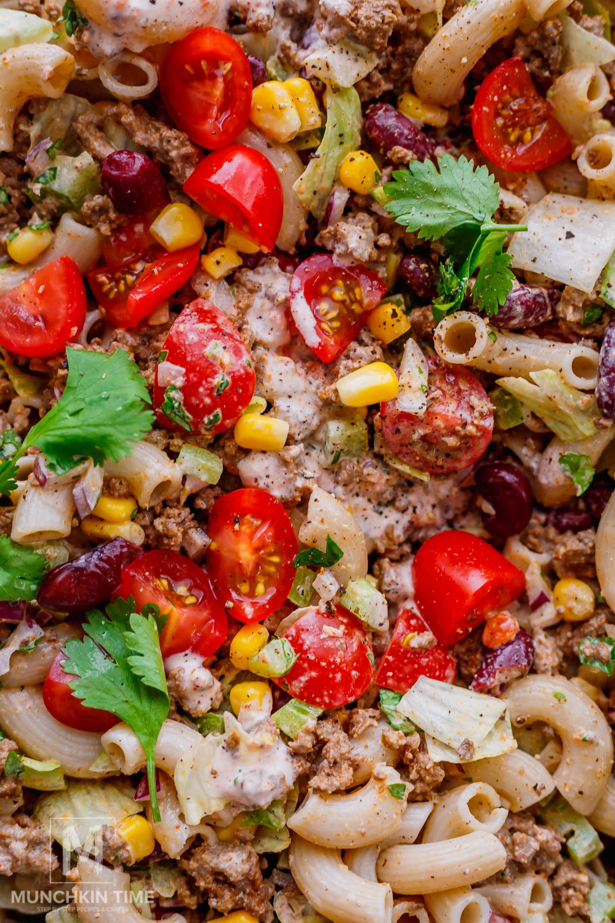 Close-up of a pasta salad with macaroni, ground meat, kidney beans, cherry tomatoes, corn, lettuce, cilantro, and a creamy dressing.