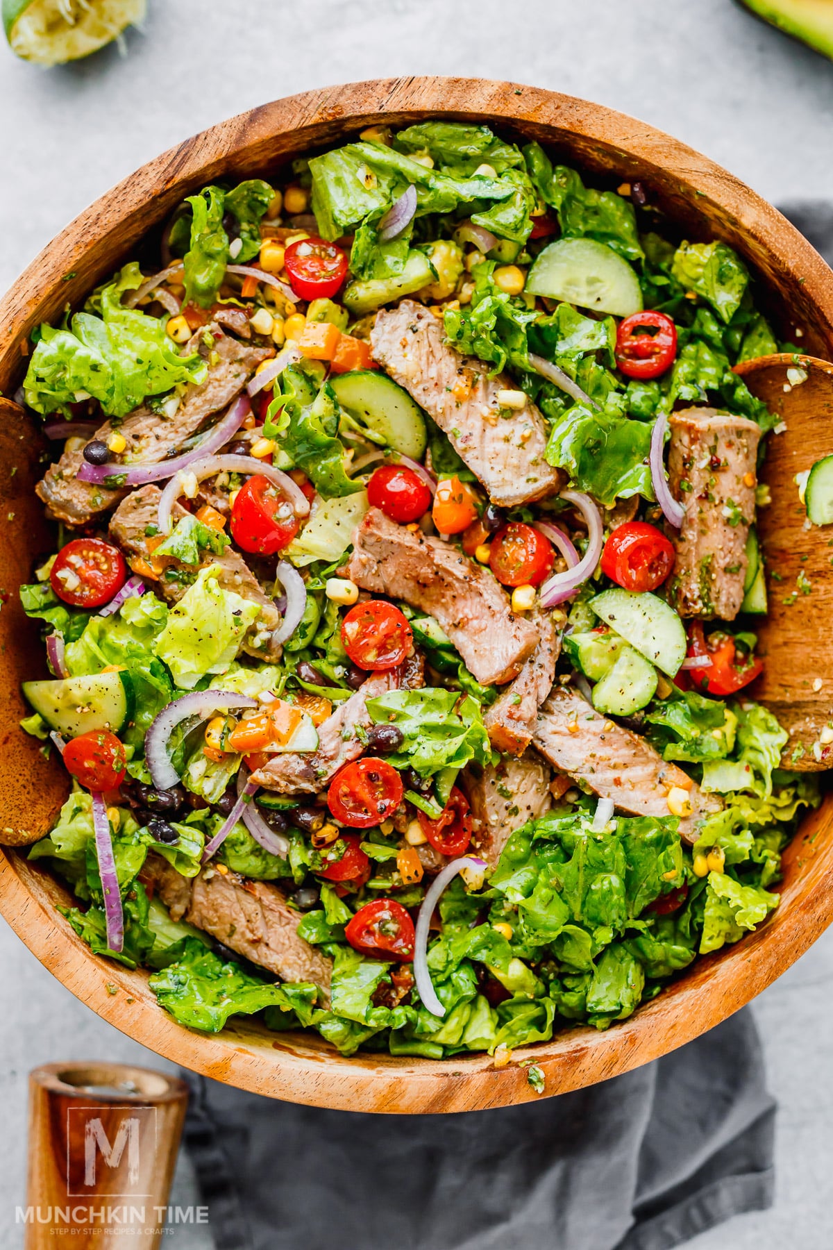 A wooden bowl filled with salad containing grilled steak slices, lettuce, cherry tomatoes, cucumbers, red onion, corn, and avocado.