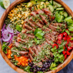 A wooden bowl filled with sliced grilled steak, cherry tomatoes, black beans, corn, avocado, red onion, bell pepper, lettuce, and herbs, arranged in sections.
