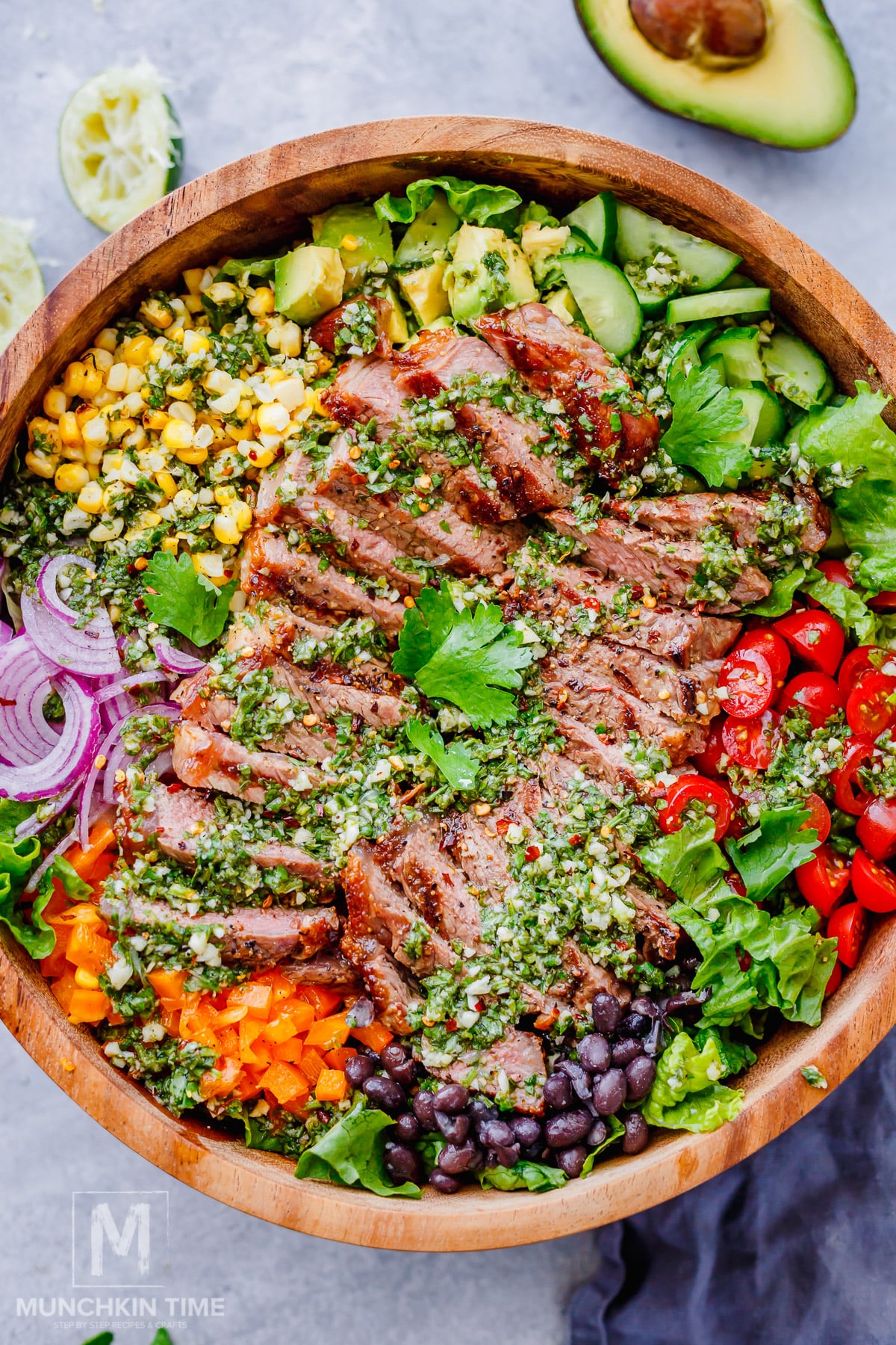 A wooden bowl filled with sliced steak, corn, avocado, cucumber, cherry tomatoes, black beans, red onion, bell pepper, greens, and chimichurri sauce.