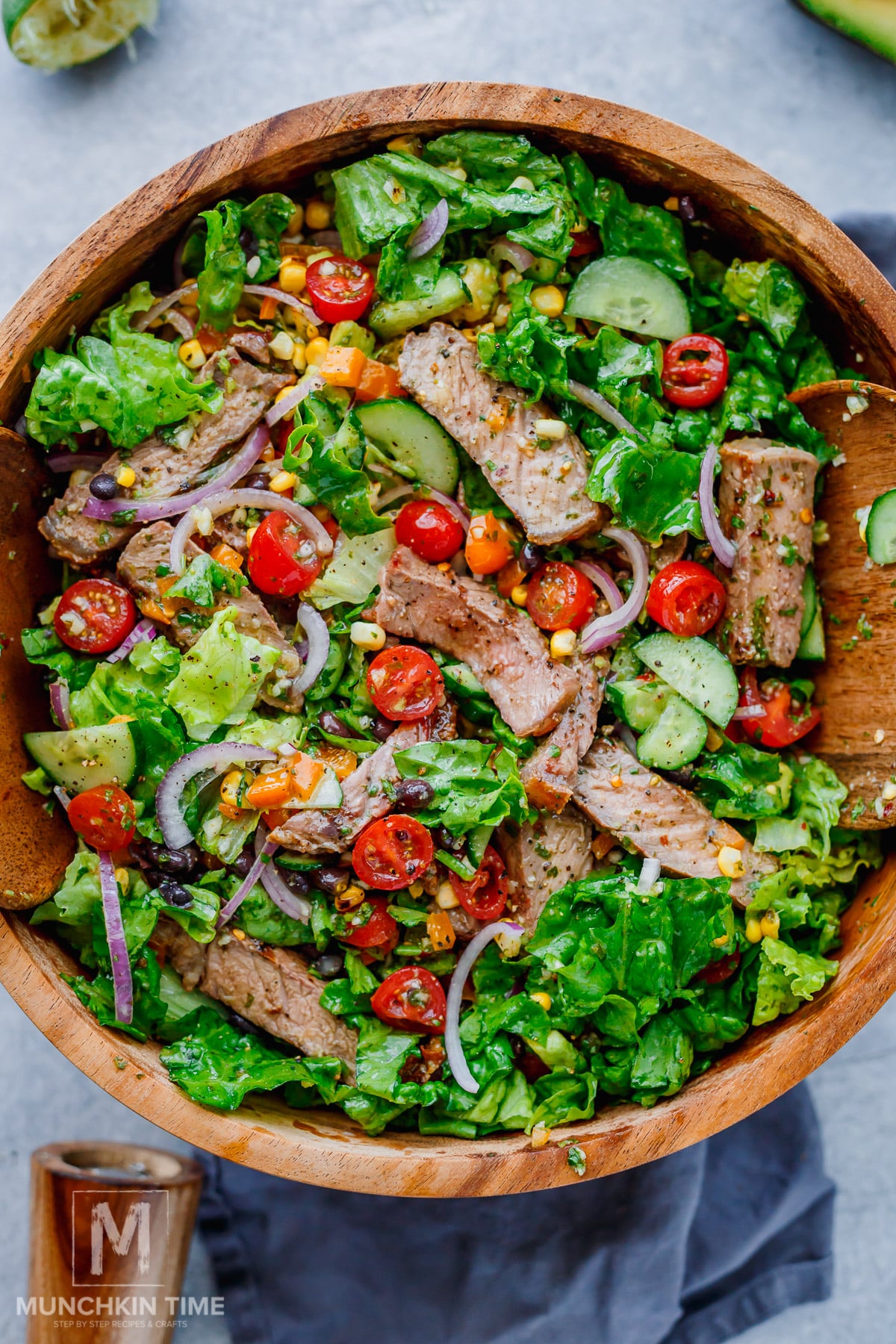 A wooden bowl filled with salad containing lettuce, grilled steak strips, cherry tomatoes, red onions, cucumbers, corn, and herbs.
