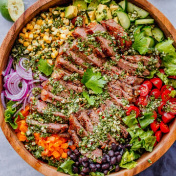 A wooden bowl filled with sliced grilled steak, cherry tomatoes, black beans, corn, avocado, red onion, bell pepper, lettuce, and herbs, arranged in sections.