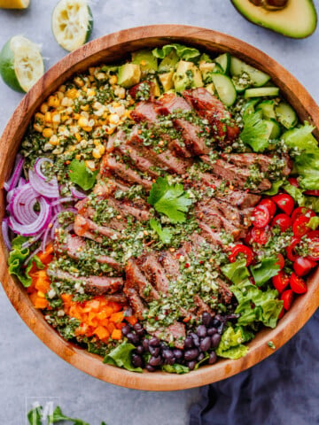A wooden bowl filled with sliced grilled steak, cherry tomatoes, black beans, corn, avocado, red onion, bell pepper, lettuce, and herbs, arranged in sections.